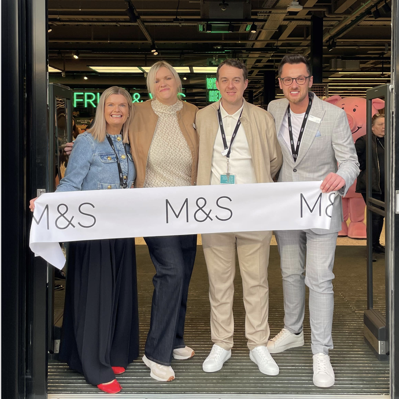 Four people standing in the doorway of a grocery store holding a ribbon that reads 'M&S'