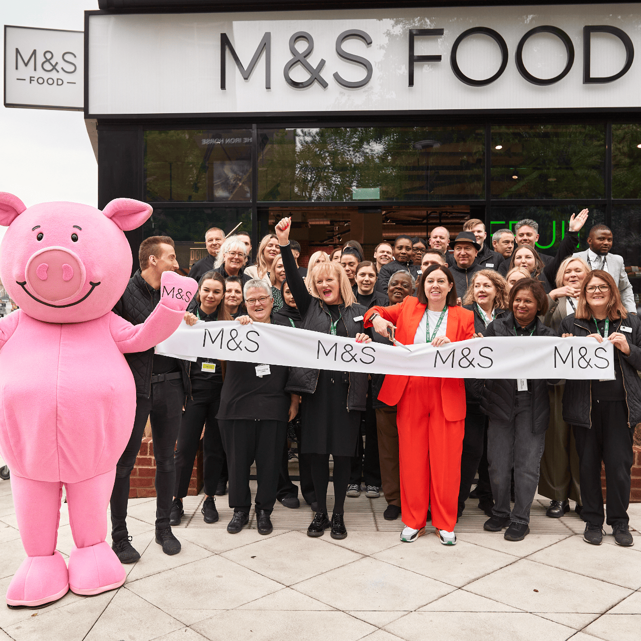 A group of people standing outside an M&S Food store with a pink pig mascot