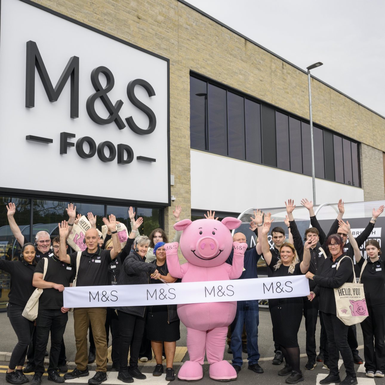 A group of people standing outside an M&S Food store with a pink pig mascott